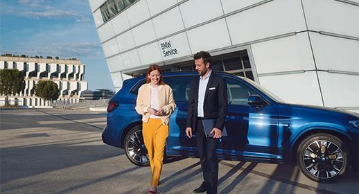 Frau und Mann gehen lächelnd vor einem BMW Service Gebäude mit einem blauen BMW SUV.
