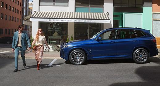 Paar läuft Hand in Hand an einem blauen BMW SUV vor einem Geschäft vorbei.